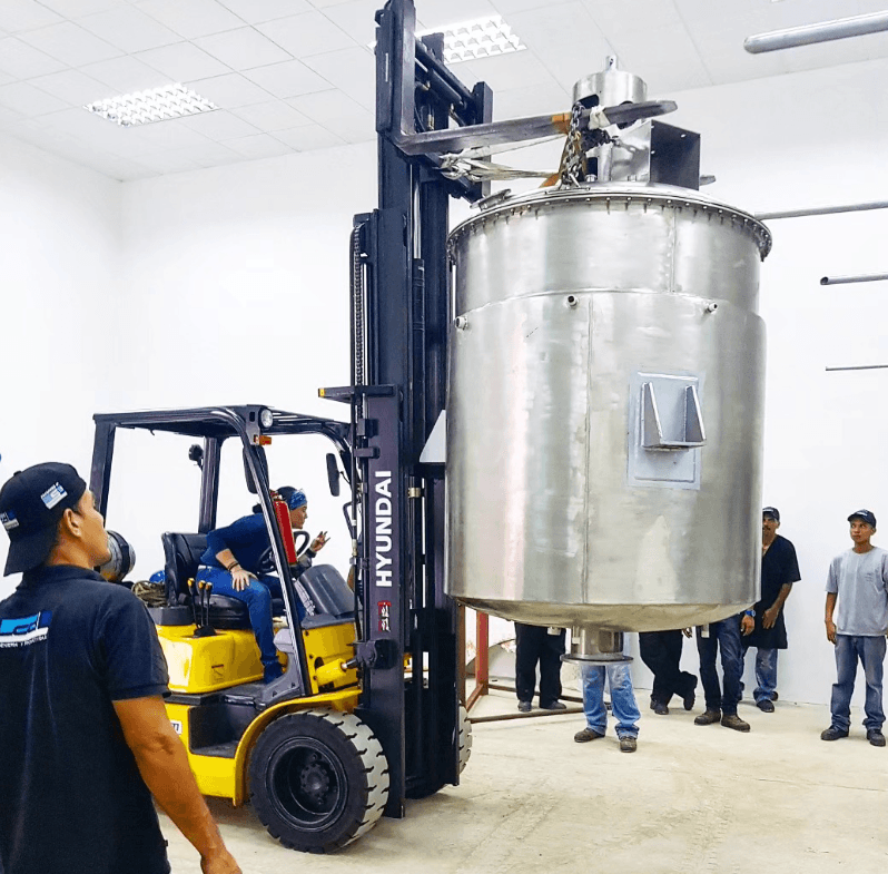Montacargas amarillo elevando un gran tanque de acero inoxidable en un almacén con trabajadores.
