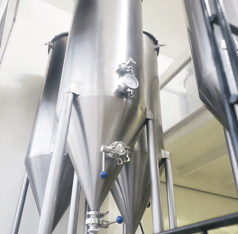 Tanques de fermentación cónicos de acero inoxidable con manómetros y válvulas en una cervecería.