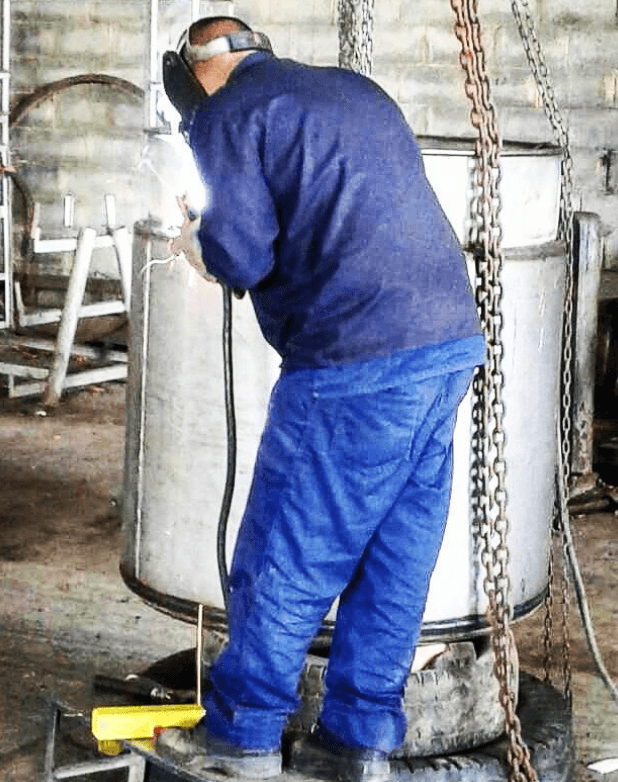 Soldador con máscara protectora y ropa azul soldando un gran cilindro metálico en un taller.