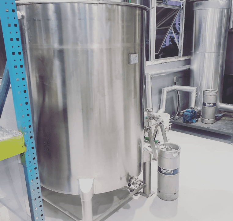 Gran tanque de acero inoxidable para elaboración de cerveza junto a un barril metálico.