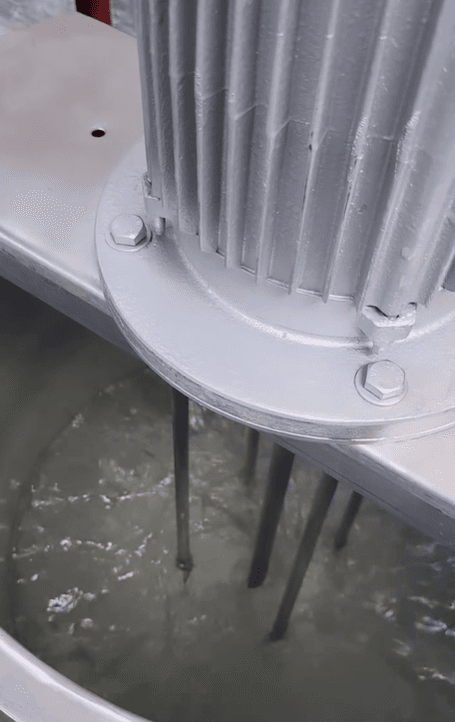 Motor industrial agitando una mezcla líquida gris con varillas metálicas dentro de un tanque metálico.