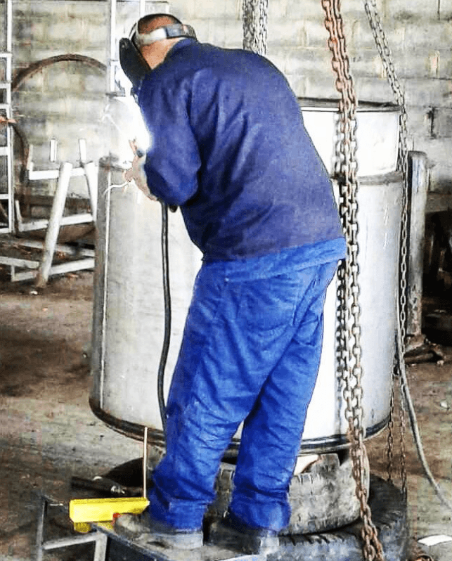 Soldador con traje azul trabajando en un gran tanque metálico cilíndrico en un taller.