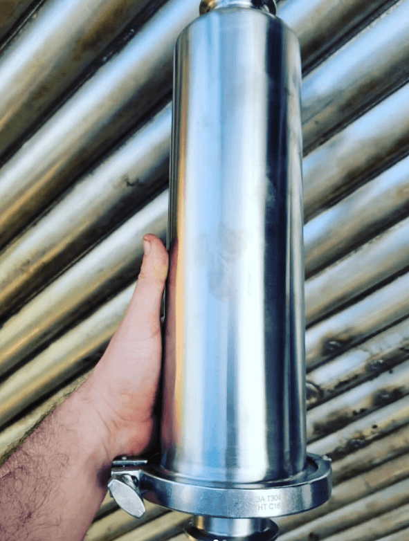 Mano sujetando una pieza cilíndrica de acero inoxidable pulido ante un fondo de tuberías industriales.