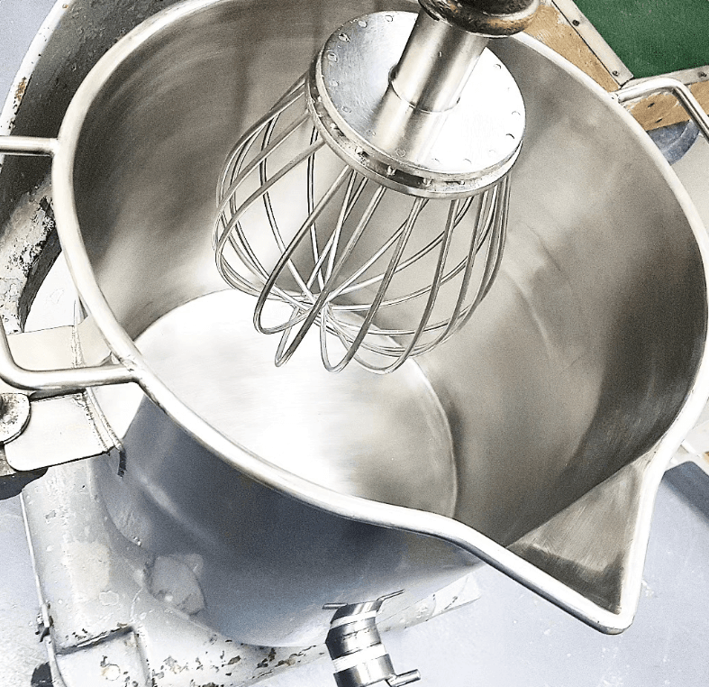 Batidora industrial de acero inoxidable con un gran batidor de varillas dentro de un tazón vacío.