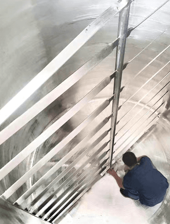 Operario dentro de un tanque cilíndrico de acero inoxidable ajustando las aspas de un agitador.